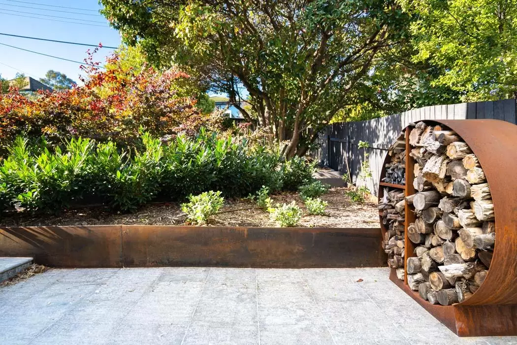 Round Corten steel firewood store beside a granite paved area and garden bed